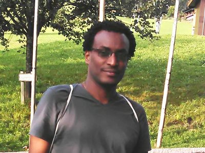 A man wearing sunglasses is standing on a street and posing for the picture, smiling into the camera. You see fields, trees, a small building and what looks like parts of a mountain or hill in the background. 