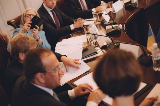 Großer, runder Sitzungstisch in einem Konferenzraum mit zahlreichen Personen, die da sitzen.