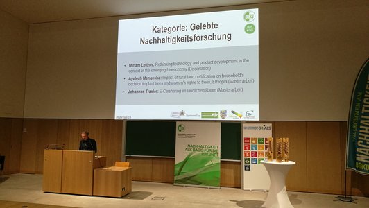 A man giving a speech from behind a speaker's desk on the stage of a large lecture hall. A power point presentation is projected onto the wall behind him, there are two roll-ups on the topic of "sustainability".