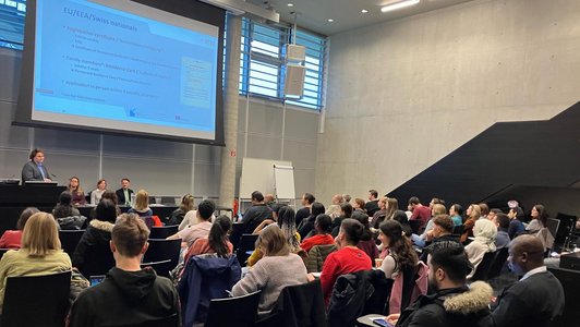 Ein Vortrag in einem gefüllten Hörsaal. Vier Personen sind auf der Bühne. Drei sitzen während ein Mann hinter dem Podium steht und ins Mikrofon redet.