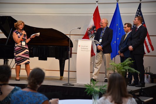 Eine Frau moderiert auf einer Bühne, daneben stehen drei Männer hinter einem Podium und vor der österreichischen, amerikanischen und der EU Flagge.