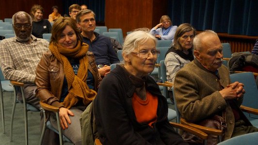 Several people as part of an audience, sitting on blue chairs.