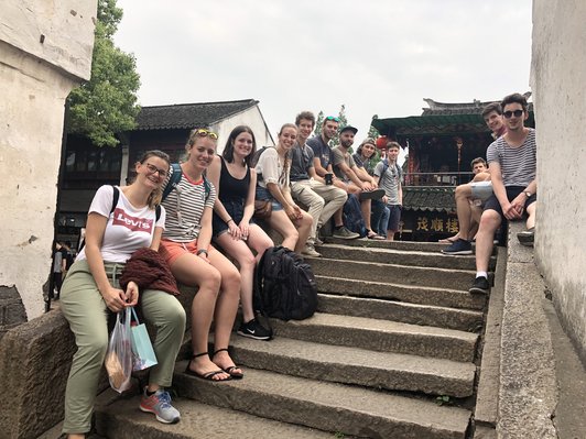  Students sit on the stone railing of a staircase in front of a temple and look into the camera