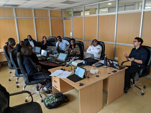 Group of people sitting around a table with notebooks on it and looking at one talking man