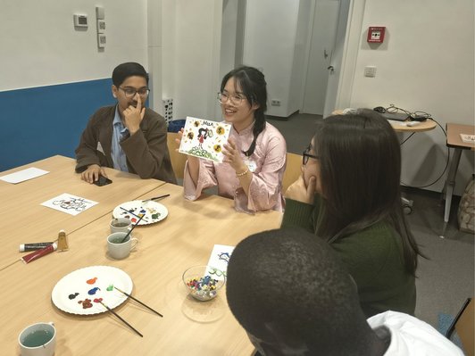 Photo of a scholarship holder showing the picture that she painted to other participants, all of them sitting around a table