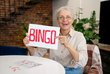 Seniorin mit Bingo-Schild, lächelnd am Tisch