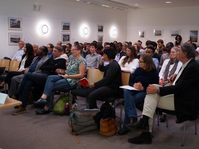 A man is giving a speech in front of an audience of approximately 50 people.