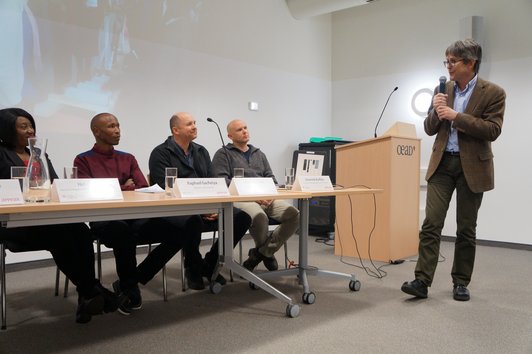 Four people are sitting at a desk with name plates, glasses of water and sheets of paper on it while one person is standing, holding a microphone and speaking to them.