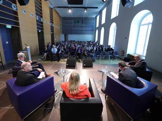 Die fünf Vertreter sitzt vor einem vollen Saal an Zuhörern, vor ihnen je zwei kleine tische mit Getränken. Jeder von ihnen hat einen Block in der Hand und eien Stift parat.