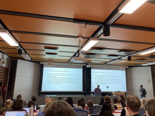 Frau mit Mikrofon an Pult, im Hintergrund Präsentation, Publikum im Vordergrund