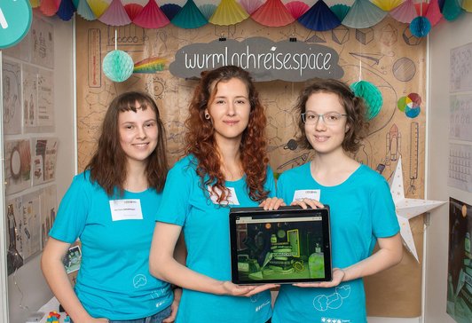 Three young girls stand in front of a kind of presentation stand at the event.