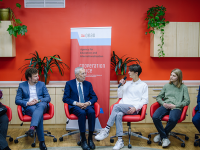 Austrian Federal President Alexander Van der Bellen sits with students and political representatives in a circle of chairs in front of a red-painted wall