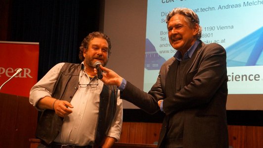 Andreas Obrecht is interviewing Andreas Melcher on the podium of an auditorium, both are smiling. 