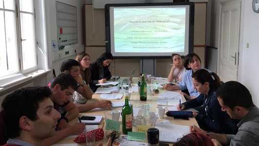 Group of people sitting at a big table in front of a presentation and looking at something else