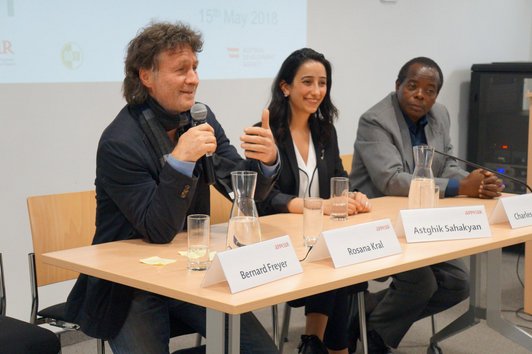 Two men and one smilling woman sitting in a row and one man is talking into a microphone