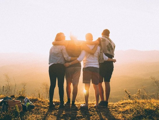 Vier Personen legen ihre Arme um die Schultern beziehungsweise um die Person neben ihnen. Sie stehen am Gipfel eines Hügels und betrachten den Sonnenuntergang.