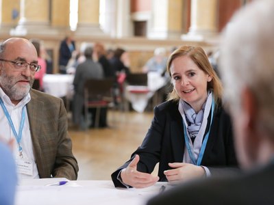 ein Mann und eine Frau sitzen an einem Tisch, die Frau spricht mit einem anderen Mann, der ihr gegenübersitzt und nur von hinten zu sehen ist, im Hintergrund weitere Personen, die an Tischen sitzen oder stehen