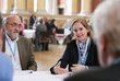 ein Mann und eine Frau sitzen an einem Tisch, die Frau spricht mit einem anderen Mann, der ihr gegenübersitzt und nur von hinten zu sehen ist, im Hintergrund weitere Personen, die an Tischen sitzen oder stehen