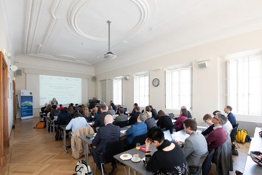 Viele Personen sitzen an Tischreihen in einem Saal und schauen nach vorne zu einer Frau, die ein Mikrophon in der Hand hält und zum Publikum spricht