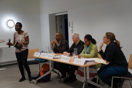 Four people are sitting at a table with name plates, glasses of water and sheets of paper on it while one person is standing, holding a microphone and a notebook in her hands, speaking.