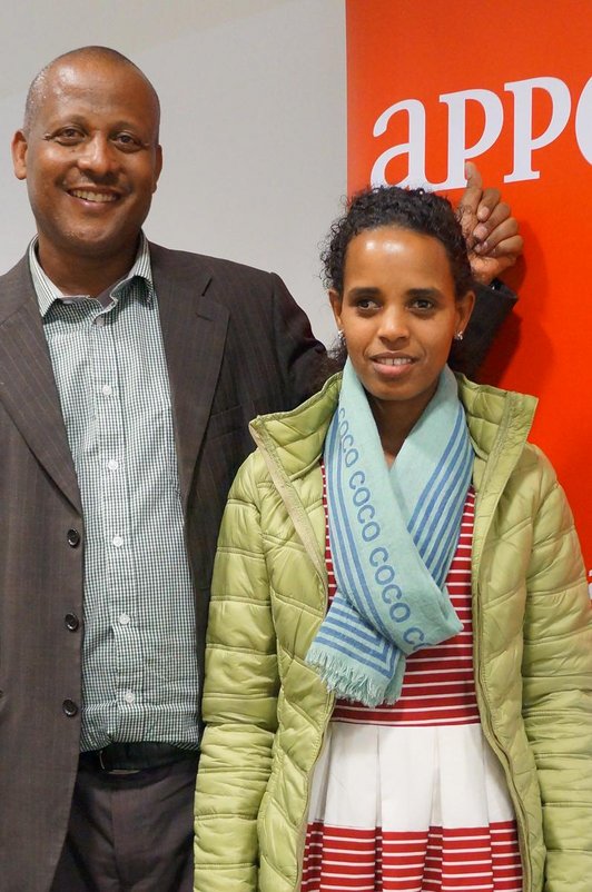 Woman and man standing side by side in front of an red APPEAR poster