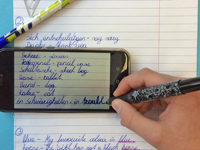  You can see a hand with a pen as well as a cell phone and a notebook. The hand writes vocabulary in English and German.