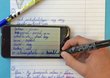  You can see a hand with a pen as well as a cell phone and a notebook. The hand writes vocabulary in English and German.