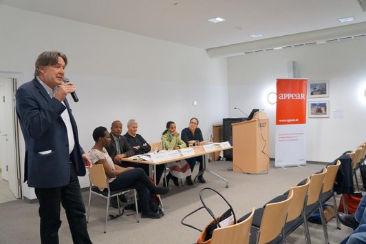 One person is giving a speech in front of an audience. In the background, five people are sitting at a table with name plates, glasses of water and sheets of paper on it.