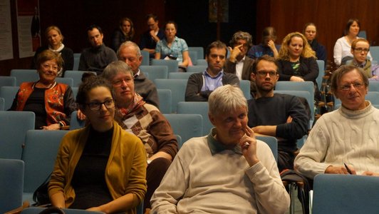 The picture shows part of an audience that is sitting on blue chairs.