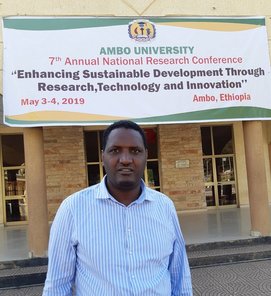 Geda Oncho standing in front of Ambo University, with a big poster in the background