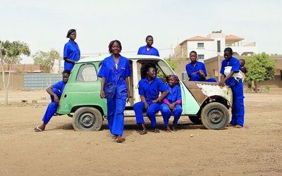 Filmsujet von Ouagagirls: acht Frauen in blauen Mechanikeruniformen stehen und sitzen rund um ein grünes Auto.