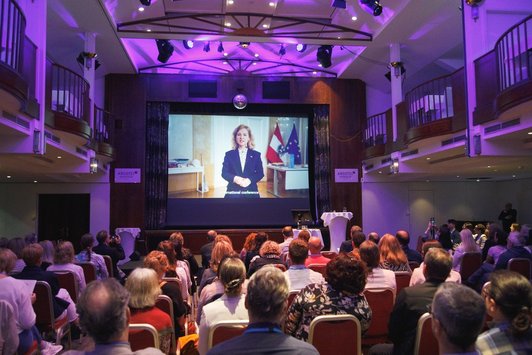 Eine Videobotschaft von BM Holzleitner wird vor den Teilnehmenden der Konferenz gezeigt.