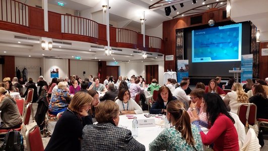 In einem Konferenzraum sitzen mehrere Personen an runden Tischen und sprechen miteinander, im Hintergrund ist der Titel der Veranstaltung auf eine Leinwand projiziert