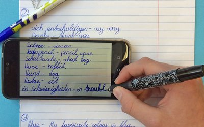 Zu sehen ist eine Hand mit einem Stift sowie ein Handy und ein Heft. Die Hand schreibt Vokabel auf englisch und deutsch. 