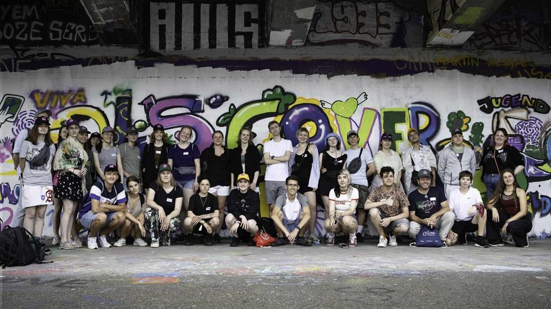 Eine Gruppe von über 30 jungen Menschen steht vor einer Wand, die mit Graffiti besprüht wurde