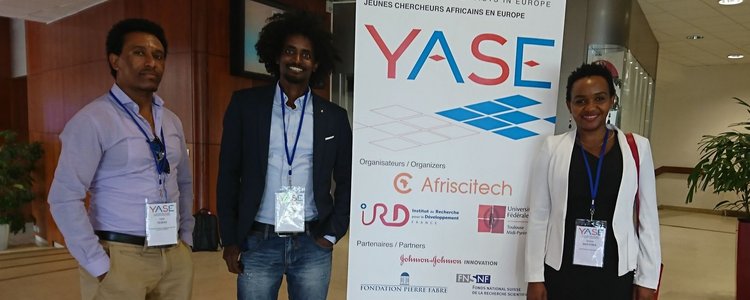One woman and two men with badges on lanyards around their necks, standing next to a roll-up announcing the "YASE, Young African Scientists in Europe" on 6 July 2018 