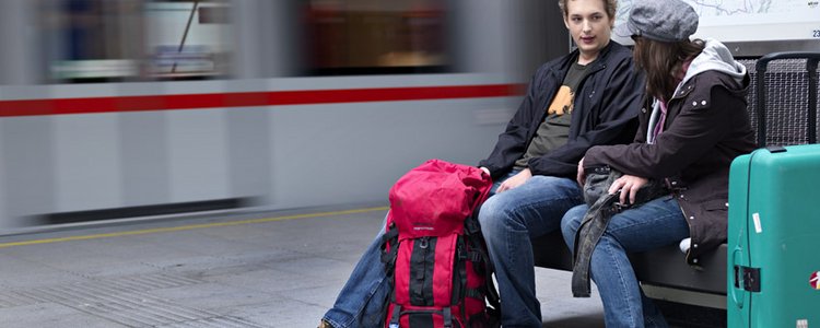 Zwei jugendliche Burschen sitzen auf einer Bank an einem Bahnsteig mit Rucksack und Koffer, im Hintergrund ein fahrender Zug.
