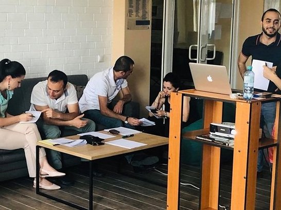 Some people sitting on a couch and some standing behind a desk with notebook on it and talking to each other