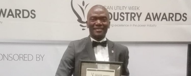 A man who has just received the African Utility Week Industry Award, standing in front of a large roll-up announcing the event.