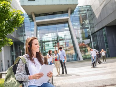 Eine junge Frau mit Rucksack steht vor einem modernen Glasgebäude.