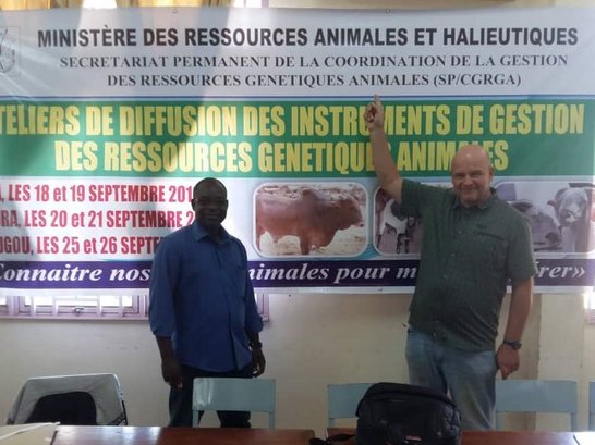 Two man smiling and standing in front of a big poster of the ministry, while one man is ponting at the poster