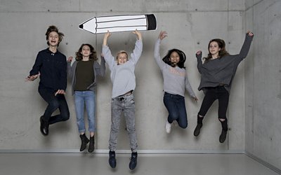 Fünf Kinder springen in die Höhe, einer hält einen großen Bleistift aus Karton in den Händen.