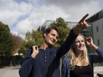 Ein junger Mann und eine junge Frau gehen nebeneinander. Er zeigt mit der ausgestreckten Hand auf etwas. 