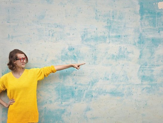 Das Bild zeigt eine Frau im gelben Pulli, die auf etwas zeigt, das nicht im Bild zu sehen ist