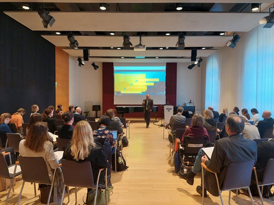 Ein Vortragender des Dialogs zur hochschulischen Lehre steht vor einer Wand mit einer Präsentation. Das Publikum ist im Hintergrund zu sehen, mit dem Rücken zur Kamera, und hört der Präsentation zu.