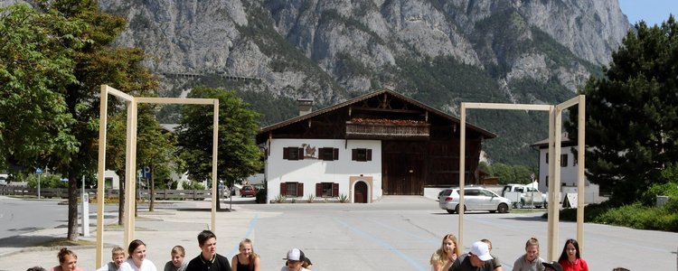Berge im Hintergrund, davor ein Haus und ein Parkplatz auf dem Gruppen von Kindern sitzen