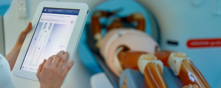 Eine medizinische Trainingspuppe liegt auf einem radiologischen Gerät, das von den Händen einer ansonsten nicht sichtbaren Person über ein Tablet bedient wird.