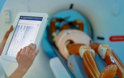 Eine medizinische Trainingspuppe liegt auf einem radiologischen Gerät, das von den Händen einer ansonsten nicht sichtbaren Person über ein Tablet bedient wird.