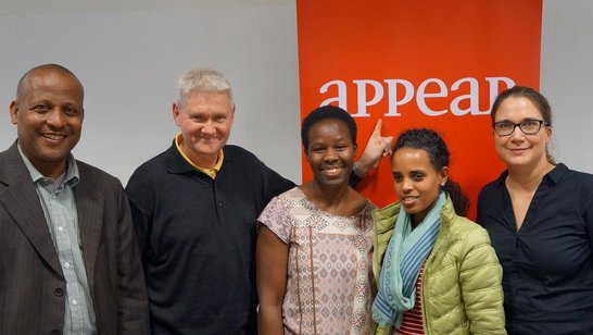 Five people are standing in front of a roll-up, one of them is pointing his finger at it. 