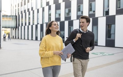 Eine Studentin und ein Student gehen nebeneinander vor einem Gebäude. 